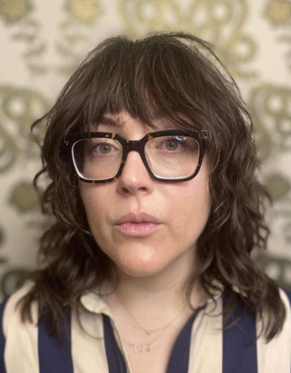 A person with a dark brown shag haircut and thick black glasses looks calmly at the camera. They are wearing a blue and white striped shirt and standing in front of green patterned wallpaper.