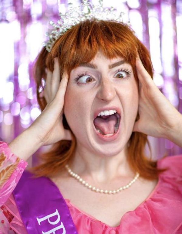 A young woman with red hair wearing a prom dress, crown, and sash is screaming comedically.