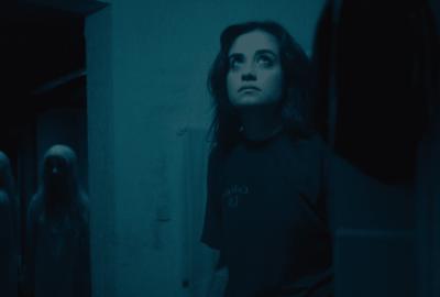 A young woman stands in a bathroom, looking upward. Behind her, barely visible in the doorway, a ghostly figure with long white hair and a pale dress lurks in the shadows.