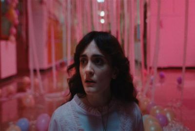 A brunette teen girl in a nightgown looks frightened in an eerie hallway surrounded by pink lighting and prom old balloons and streamers.