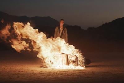 Irene stands in front of a burning man in a chair