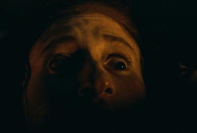 The upper half of a young woman's face lit by dim candlelight. She appears frightened.