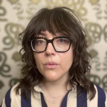 A person with a dark brown shag haircut and thick black glasses looks calmly at the camera. They are wearing a blue and white striped shirt and standing in front of green patterned wallpaper.