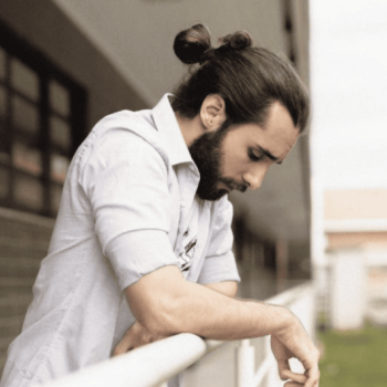 A bearded man with long hair in a bun leans on a railing, looking down thoughtfully.